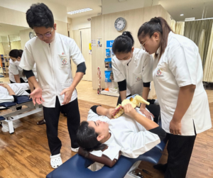 仰向けで寝ながら背骨の模型をもっているセラピストそれを見てる二人のセラピスト、立って両手を肩幅くらいに広げているセラピスト