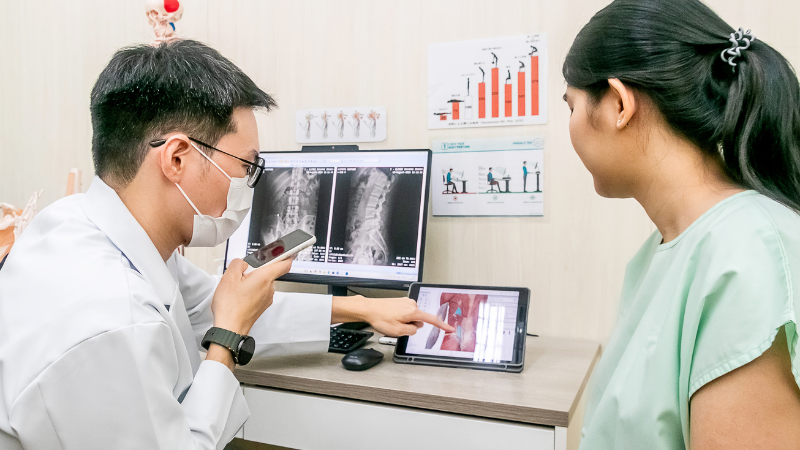 医師がAI音声翻訳機をもって口元にあてている、解剖アプリのスクリーンを指さしている、それをみる患者