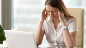 痛そうに頭のこめかみを抑えている女性目の前にはパソコンとメガネがおいてある