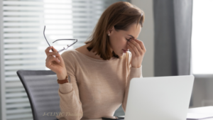 パソコンの前で左手で目頭を押さえ、右手にメガネをもっている女性