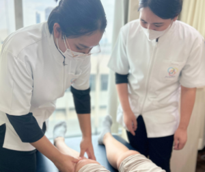 仰向けになった人の膝を触るセラピストとそれを見ているセラピスト