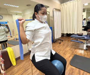 セラピストが椅子に座って、膝の間にソフトボールを挟んで、セラバンドを両手にもって首の後ろ側にあげている
