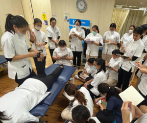 うつ伏せになった人の右足を持ち上げているセラピスト、それをみている大勢のセラピスト